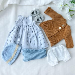 May include: A collection of baby clothes, including a light blue and white striped dress, a brown knitted cardigan with two buttons, a blue knitted hat, a blue knitted neck warmer, white bloomers, and gray knitted baby shoes.