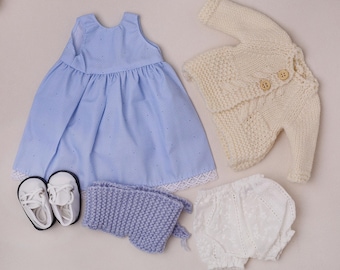 May include: A baby doll clothing set featuring a light blue dress with white lace trim, a cream-colored knitted sweater, white bloomers, a lavender bonnet, and white shoes with black soles. The dress has a polka dot pattern.