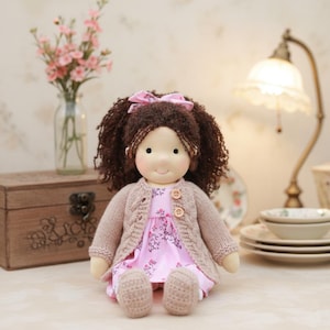 May include: A handmade doll with brown curly hair, wearing a pink floral dress and a beige knitted cardigan. The doll has a pink bow in her hair and is sitting with her legs crossed. The doll is a soft toy.