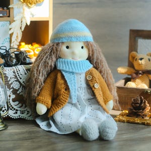 K&ouml;nnte beinhalten: Handgemachte Puppe mit langen braunen Haaren, die eine hellblaue und gelbe Strickm&uuml;tze, einen blauen Schal und eine senfgelbe Strickjacke tr&auml;gt. Die Puppe tr&auml;gt ein wei&szlig;es Kleid mit kleinen schwarzen Punkten und grauen Strickschuhen.
