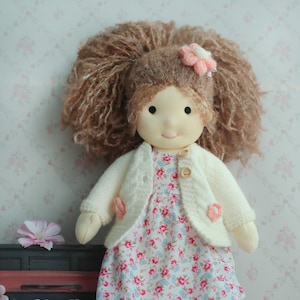 May include: A handmade doll with a soft, smiling face, wearing a floral dress and a cream-colored cardigan. The doll has brown, fluffy hair and a pink flower accent. The doll is a charming, handcrafted item.