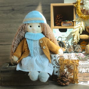 K&ouml;nnte beinhalten: Eine handgefertigte Puppe mit langen braunen Haaren, die eine hellblaue und beige Strickm&uuml;tze, einen passenden Schal und eine senfgelbe Strickjacke tr&auml;gt. Die Puppe tr&auml;gt ein wei&szlig;es Kleid und hat wei&szlig;e Schuhe. Die Puppe sitzt auf einer Holzkiste.