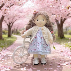 May include: A handmade doll with long, wavy brown hair, wearing a floral dress and a cream-colored cardigan. The doll is holding a white bicycle and is standing on a path lined with cherry blossom trees. The doll is wearing white shoes.