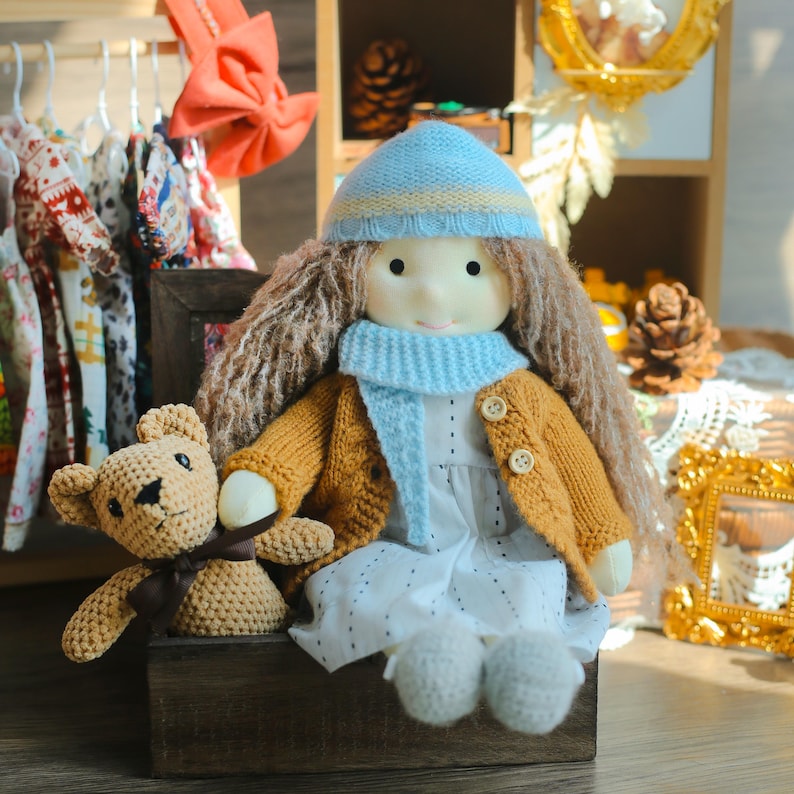 K&ouml;nnte beinhalten: Eine handgefertigte Puppe mit langen braunen Haaren, die einen blau-gelben Hut, einen blauen Schal und eine senffarbene Strickjacke tr&auml;gt. Ein kleiner geh&auml;kelter Teddyb&auml;r sitzt neben der Puppe. Die Puppe tr&auml;gt ein wei&szlig;es Kleid mit kleinen schwarzen Punkten.