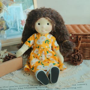 May include: A handmade doll with brown curly hair and a smiling face. The doll wears a yellow dress with a white daisy pattern and black shoes. The doll is sitting on a lace-covered surface.