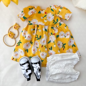 May include: A yellow baby dress with a white daisy floral pattern, short sleeves, and a round neckline. The image also shows a pair of black and white sneakers, a white lace diaper cover, and a yellow hair accessory with cartoon bear faces.