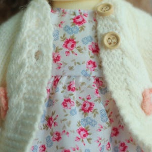 May include: Close-up of a doll's outfit featuring a cream-colored knitted cardigan with wooden buttons over a floral print dress. The dress has a white background with pink and blue floral patterns. The outfit is handmade.