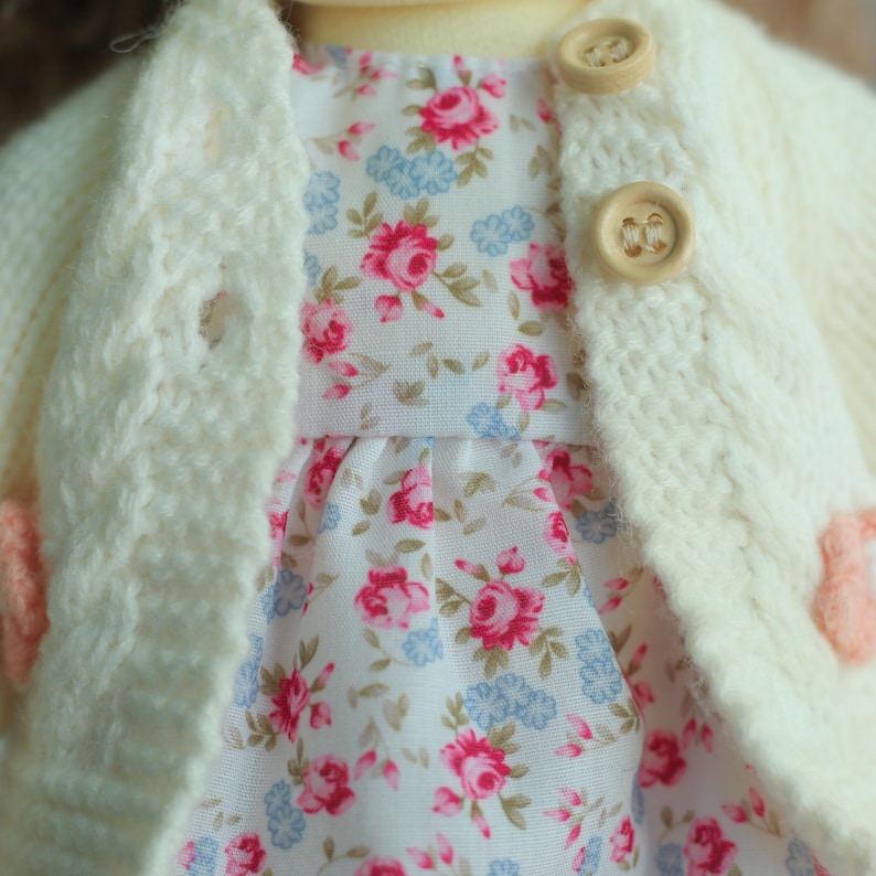 May include: Close-up of a doll's outfit featuring a cream-colored knitted cardigan with wooden buttons over a floral print dress. The dress has a white background with pink and blue floral patterns. The outfit is handmade.