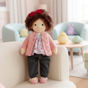 May include: A handmade doll with brown curly hair and a red bow. The doll wears a pink cable-knit cardigan, a floral print blouse, dark gray pants, and pink shoes. The doll is sitting on a cream-colored sofa.