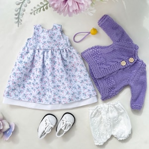 Puede incluir: Un conjunto de ropa de muñeca con un vestido con estampado floral, una rebeca de punto morado con botones de madera, pantalones cortos de encaje blancos, zapatos blancos y una goma para el pelo amarilla. El vestido tiene un dobladillo blanco.