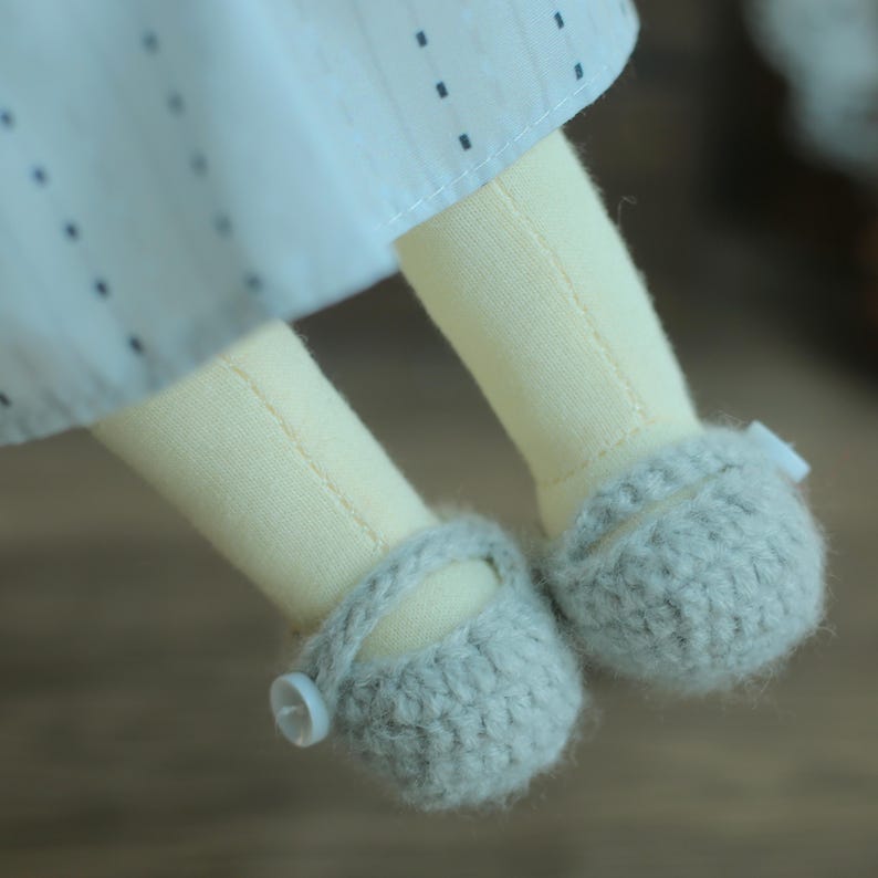 K&ouml;nnte beinhalten: Nahaufnahme der Beine und F&uuml;&szlig;e einer Puppe. Die Beine der Puppe sind aus hellgelbem Stoff, und die F&uuml;&szlig;e tragen graue Strickschuhe mit Knopfverschl&uuml;ssen. Die Puppe tr&auml;gt ein wei&szlig;es Kleid mit kleinen schwarzen Punkten.