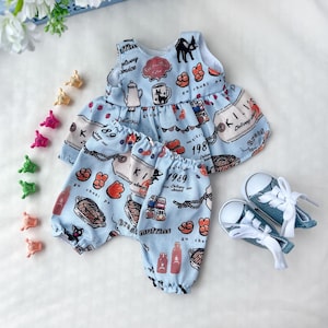 May include: A light blue doll outfit with a food-themed print, including a top and pants. The outfit features a black cat, bread, and the text "KI 1989". A pair of denim doll shoes with white laces are also included.