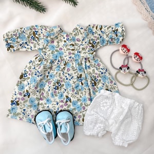 May include: A baby's floral dress with blue flowers, matching blue shoes, white lace shorts, and hair ties with cartoon character faces. The dress has short sleeves and a gathered skirt. The shoes have white laces.