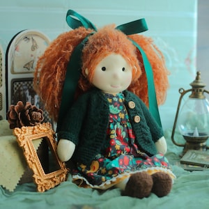 Puede incluir: Muñeca hecha a mano con cabello de hilo rojo atado con cinta verde. La muñeca lleva una rebeca de punto verde oscuro sobre un vestido con estampado floral. La muñeca tiene zapatos con botones marrones y una cara sonriente.