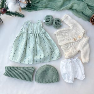 May include: A baby clothing set featuring a light green dress with black vertical stripes, a white knitted sweater and pants, a green knitted hat, a green knitted neck warmer, white bloomers, and green knitted shoes.