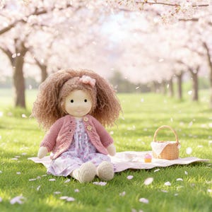 Puede incluir: Una muñeca hecha a mano con cabello castaño rizado, vestida con una rebeca rosa y un vestido floral, está sentada sobre una manta de picnic. Una pequeña cesta de mimbre, una taza de té y una magdalena están cerca sobre la hierba, con cerezos en flor al fondo.