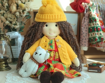 May include: A handmade doll with long brown hair, wearing a yellow knit hat and sweater over a patterned dress. The doll holds a small white teddy bear. The doll's outfit includes a red, yellow, and green patterned dress. The doll is sitting on a white lace doily.