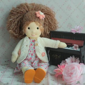 May include: A handmade doll with a large brown yarn hairstyle and a pink flower hair accessory. The doll wears a floral print dress, a cream-colored cardigan, and orange shoes. A miniature black piano and pink flowers are in the background.