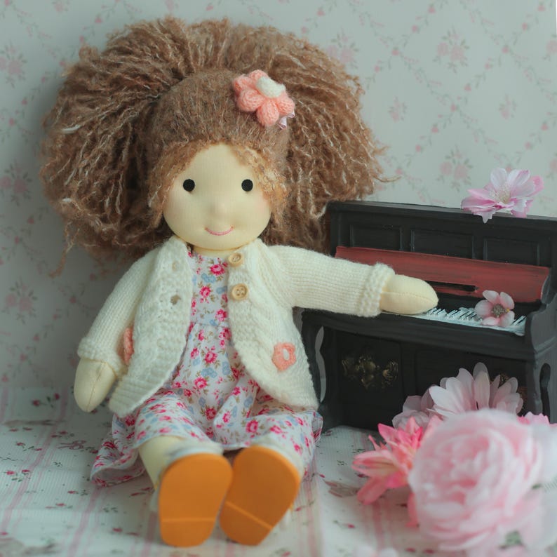 May include: A handmade doll with a large brown yarn hairstyle and a pink flower hair accessory. The doll wears a floral print dress, a cream-colored cardigan, and orange shoes. A miniature black piano and pink flowers are in the background.