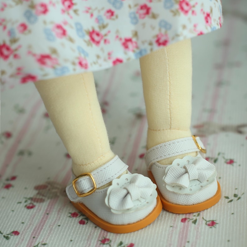 May include: Close-up of a doll's feet wearing white Mary Jane shoes with orange soles and gold buckles. Each shoe has a white bow on the toe. The doll is wearing a floral print dress.