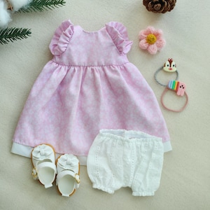 May include: A pink floral print dress with ruffled sleeves and a white hem, paired with white lace bloomers and white baby shoes. Accessories include a pink flower and a colorful hair tie.