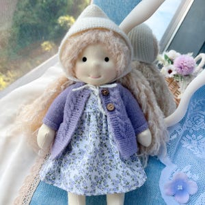 May include: A handcrafted doll with long blonde hair, wearing a white bonnet, a lavender cable-knit cardigan, and a floral print dress. The doll has a sweet, simple face and is made of soft materials. The doll is a charming example of handcrafted artistry.