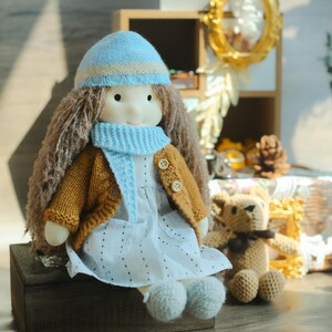 K&ouml;nnte beinhalten: Eine handgemachte Puppe mit langen braunen Haaren, die einen hellblauen Hut und Schal sowie eine senfgelbe Strickjacke &uuml;ber einem wei&szlig;en Kleid tr&auml;gt. Die Puppe sitzt auf einer Holzkiste, mit einem kleinen geh&auml;kelten Teddyb&auml;ren in der N&auml;he.