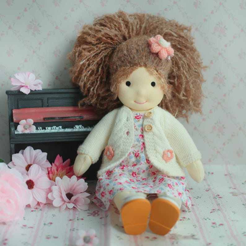 May include: A handmade doll with brown yarn hair and a pink flower hair accessory. The doll wears a floral print dress, a cream-colored cardigan with pink flower accents, and orange shoes. The doll is sitting near a miniature piano and pink flowers.