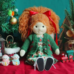May include: A handmade doll with red yarn hair tied with a red ribbon, wearing a green sweater and a patterned dress. The doll has a smiling face and is posed with Christmas decorations, including a small train and snowmen.
