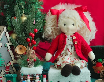 May include: A handmade doll with blonde curly hair and a red bow sits on a white platform. The doll wears a red cardigan over a Christmas-themed dress. A Christmas tree and decorative train set are in the background, creating a festive holiday scene.