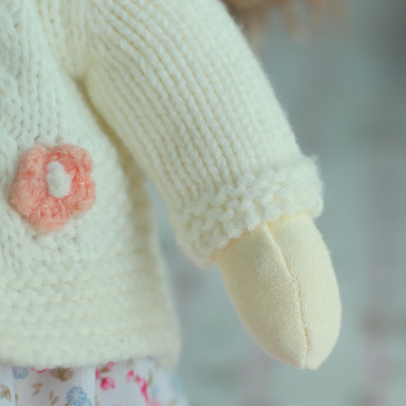 May include: Close-up of a doll's arm and part of its body. The doll wears a cream-colored knitted sweater with a pink flower applique. The doll's arm is made of a light beige fabric.