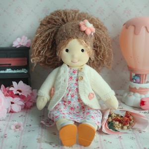 May include: A handmade doll with a soft, fabric face, brown yarn hair, and a pink flower hair accessory. The doll wears a floral print dress, a cream-colored knitted cardigan, and orange shoes. A small bouquet of dried flowers rests nearby.
