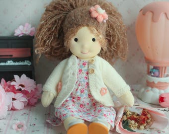 May include: A handmade doll with a soft, fabric face, brown yarn hair, and a pink flower hair accessory. The doll wears a floral print dress, a cream-colored knitted cardigan, and orange shoes. A small bouquet of dried flowers rests nearby.