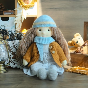 K&ouml;nnte beinhalten: Handgemachte Puppe mit langen braunen Haaren, tr&auml;gt eine hellblaue Strickm&uuml;tze und einen Schal sowie eine senfgelbe Strickjacke. Die Puppe tr&auml;gt ein wei&szlig;es Kleid und graue Schuhe. Die Puppe sitzt auf einer Holzoberfl&auml;che.