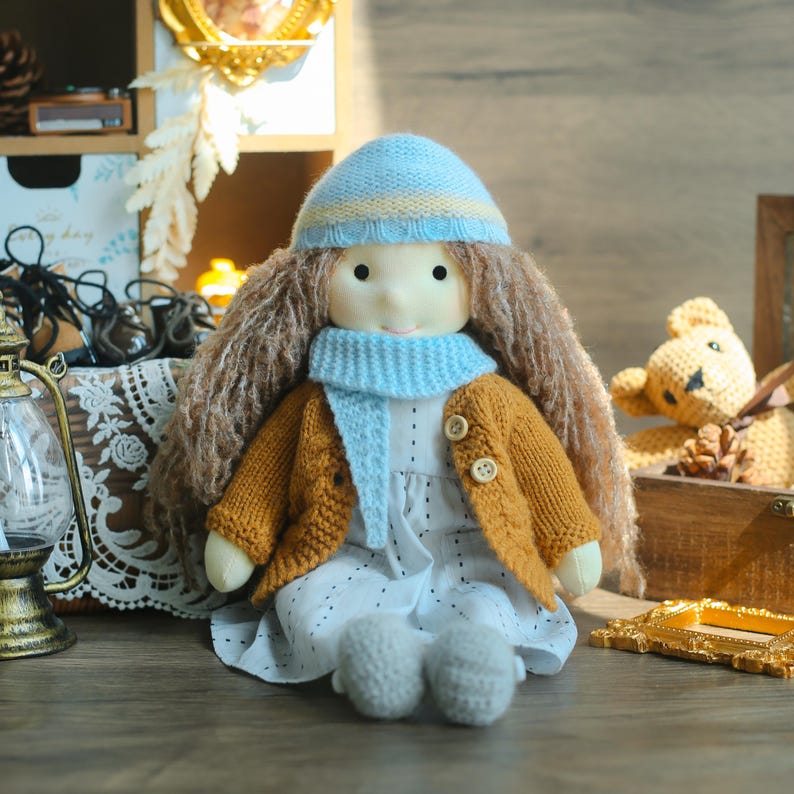 K&ouml;nnte beinhalten: Handgemachte Puppe mit langen braunen Haaren, tr&auml;gt eine hellblaue Strickm&uuml;tze und einen Schal sowie eine senfgelbe Strickjacke. Die Puppe tr&auml;gt ein wei&szlig;es Kleid und graue Schuhe. Die Puppe sitzt auf einer Holzoberfl&auml;che.