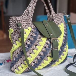 May include: A handcrafted crochet handbag with a distinctive pattern. The bag combines grey, yellow, and blue yarn, complemented by a green leather flap and strap. It features a silver buckle and a robust handle.