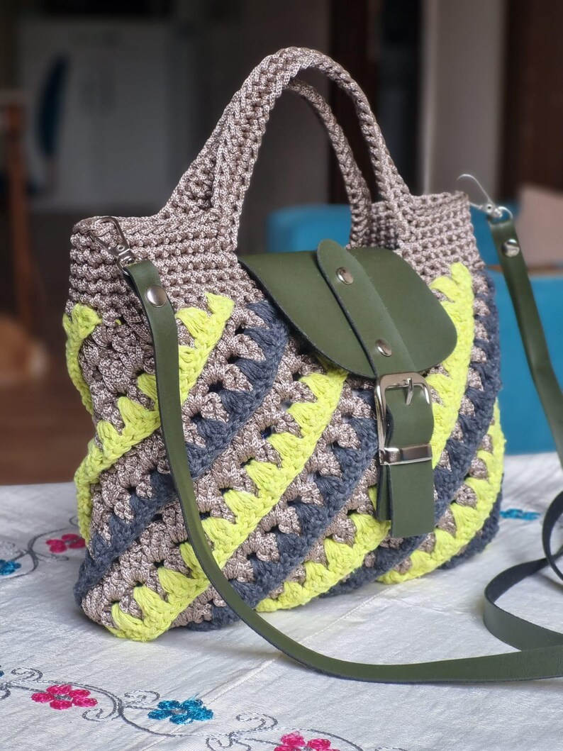 May include: A handcrafted crochet handbag with a distinctive pattern. The bag combines grey, yellow, and blue yarn, complemented by a green leather flap and strap. It features a silver buckle and a robust handle.