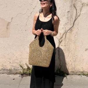 May include: A woven straw tote bag with a wide opening and two handles. The bag is a light tan colour and appears to be handmade. The bag is being held in front of a black dress.