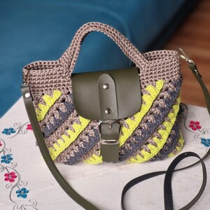 May include: A crochet handbag with a flap closure. The bag is made with grey, yellow, and brown crochet. It has a green leather flap and strap, and a silver buckle. The bag has a top handle and a long shoulder strap.