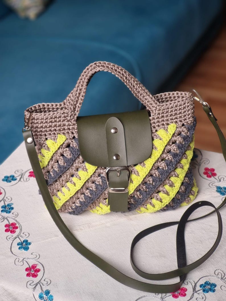 May include: A crochet handbag with a flap closure. The bag is made with grey, yellow, and brown crochet. It has a green leather flap and strap, and a silver buckle. The bag has a top handle and a long shoulder strap.