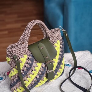 May include: A crochet handbag with a geometric pattern in grey, yellow, and blue. It has a green leather flap with a silver buckle and a matching green shoulder strap. The bag has a braided handle.