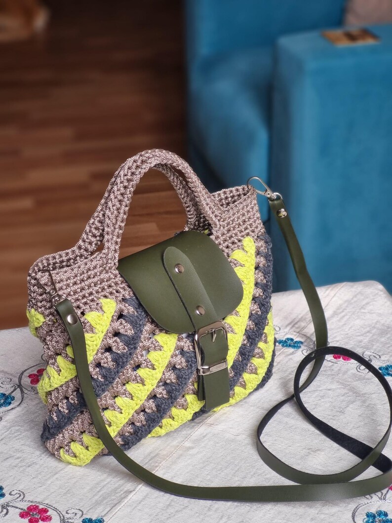 May include: A crochet handbag with a geometric pattern in grey, yellow, and blue. It has a green leather flap with a silver buckle and a matching green shoulder strap. The bag has a braided handle.
