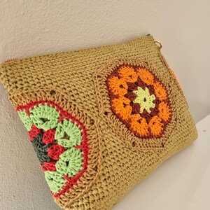 May include: A tan woven clutch bag with colourful crochet floral designs. The crochet details include orange, green, red, and yellow. The bag has a zip closure and a gold-coloured ring attached to the zip.