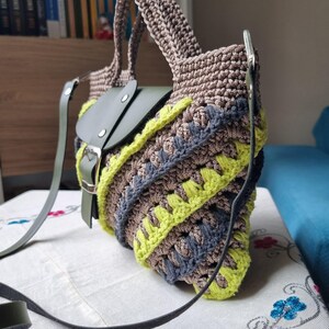 May include: A handcrafted crochet bag in shades of brown, grey, and neon yellow. It has a green leather flap and strap, a secure buckle, and a distinctive pattern. The bag's design is eye-catching and the strap is adjustable.