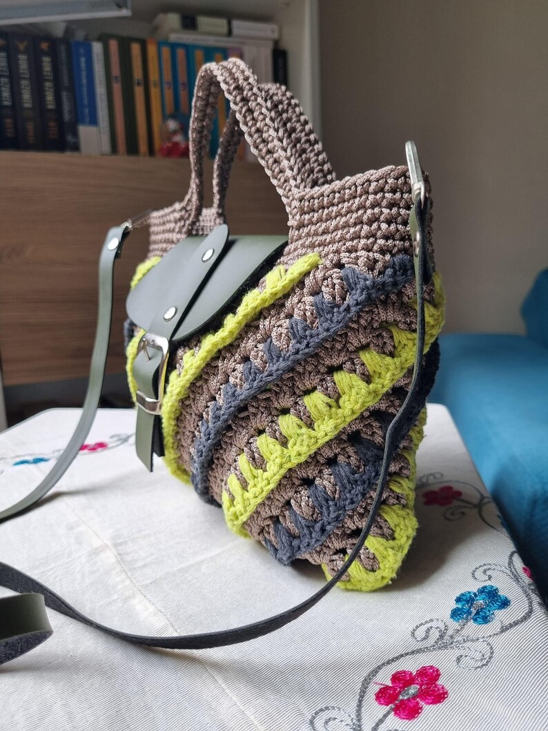 May include: A handcrafted crochet bag in shades of brown, grey, and neon yellow. It has a green leather flap and strap, a secure buckle, and a distinctive pattern. The bag's design is eye-catching and the strap is adjustable.