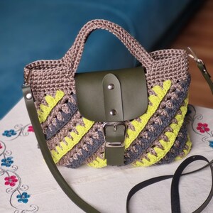 May include: A crochet handbag with a brown top and handle, a green leather flap, and a detachable green strap. The bag has a diagonal pattern of grey and yellow stripes.
