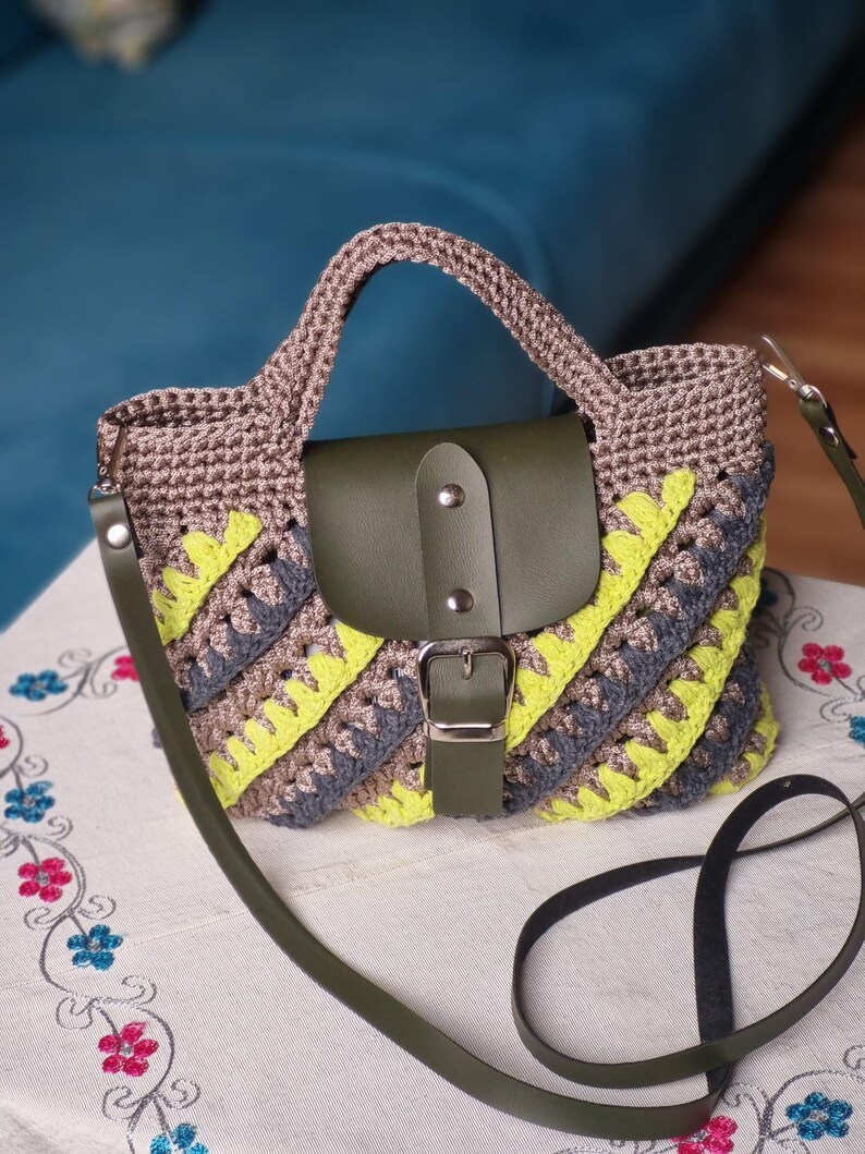 May include: A crochet handbag with a brown top and handle, a green leather flap, and a detachable green strap. The bag has a diagonal pattern of grey and yellow stripes.