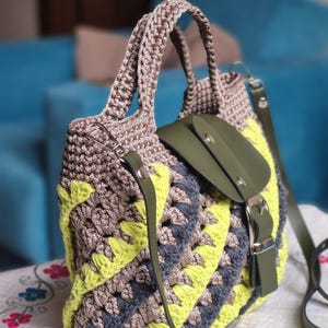 May include: A handmade crochet tote bag in shades of grey, yellow, and blue. The bag features a geometric pattern, a green leather flap with a buckle closure, and a matching shoulder strap. The handles are also crocheted.