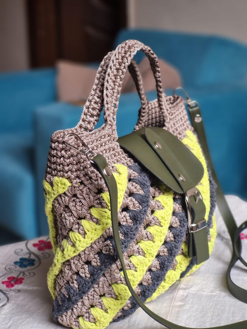 May include: A handmade crochet tote bag in shades of grey, yellow, and blue. The bag features a geometric pattern, a green leather flap with a buckle closure, and a matching shoulder strap. The handles are also crocheted.