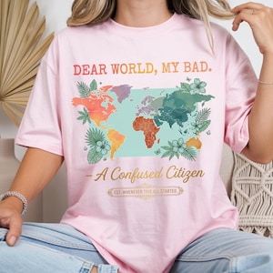 May include: Pink t-shirt with the text "DEAR WORLD, MY BAD." and "- A Confused Citizen." A colorful world map graphic is featured on the front, with floral and leaf accents. The shirt also has the text "EST. WHENEVER THIS ALL STARTED."